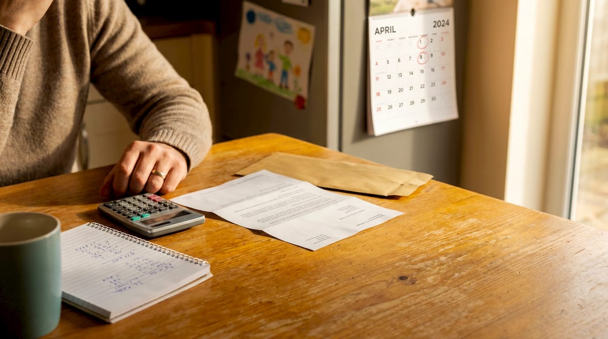 A parent at a UK kitchen table with an opened benefits uprating letter, a calculator and a wall calendar turned to April. AI-generated illustration.