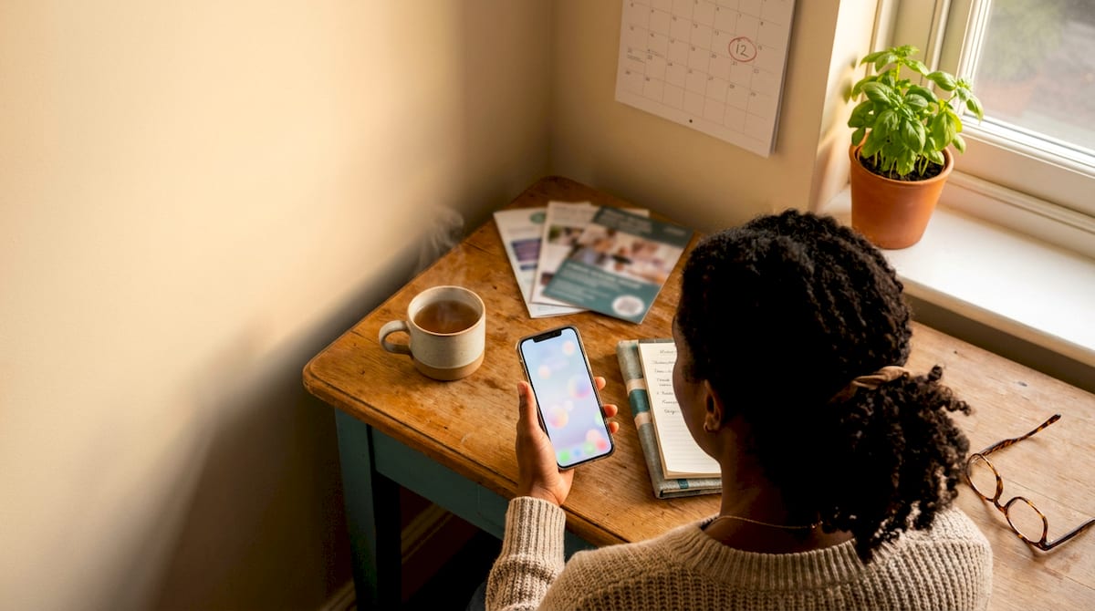 A parent at a small home desk checking what support they can claim on their phone, with notes and a calendar nearby. AI-generated illustration.