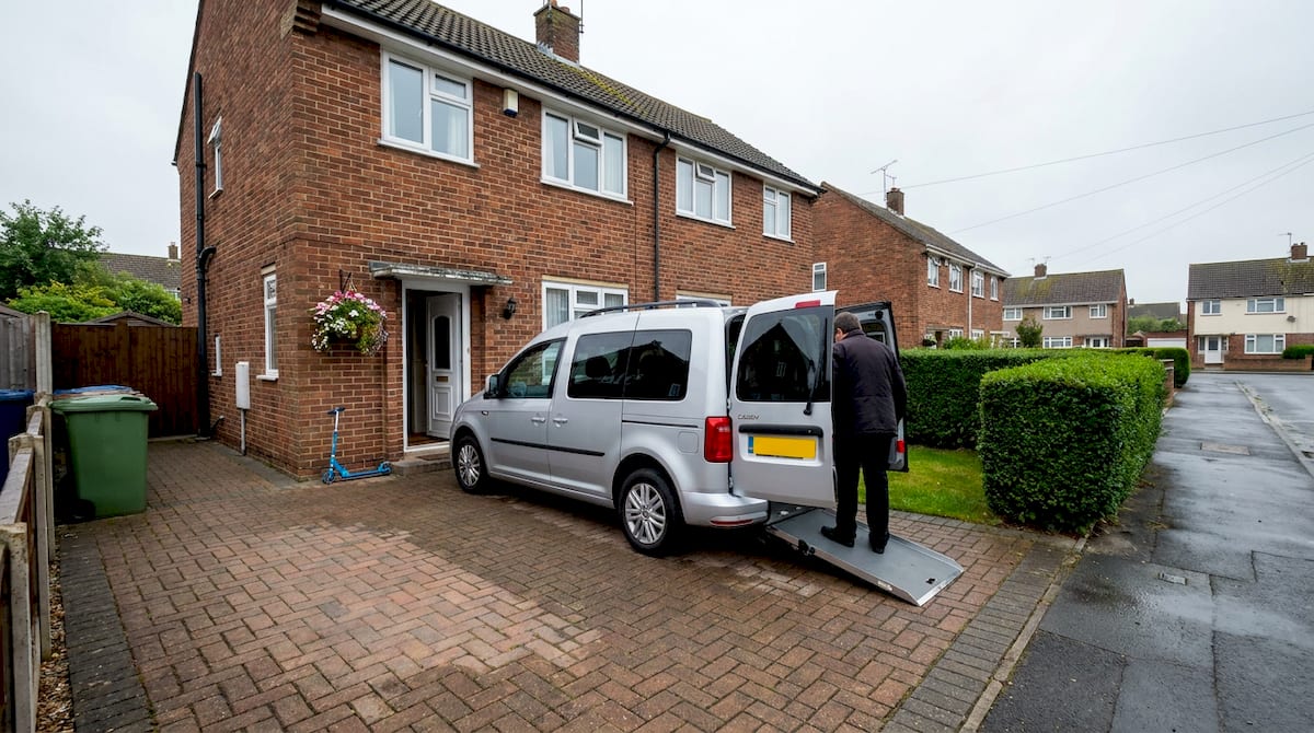 A wheelchair-accessible vehicle with its rear ramp lowered on a UK driveway outside a brick semi-detached house. AI-generated illustration.