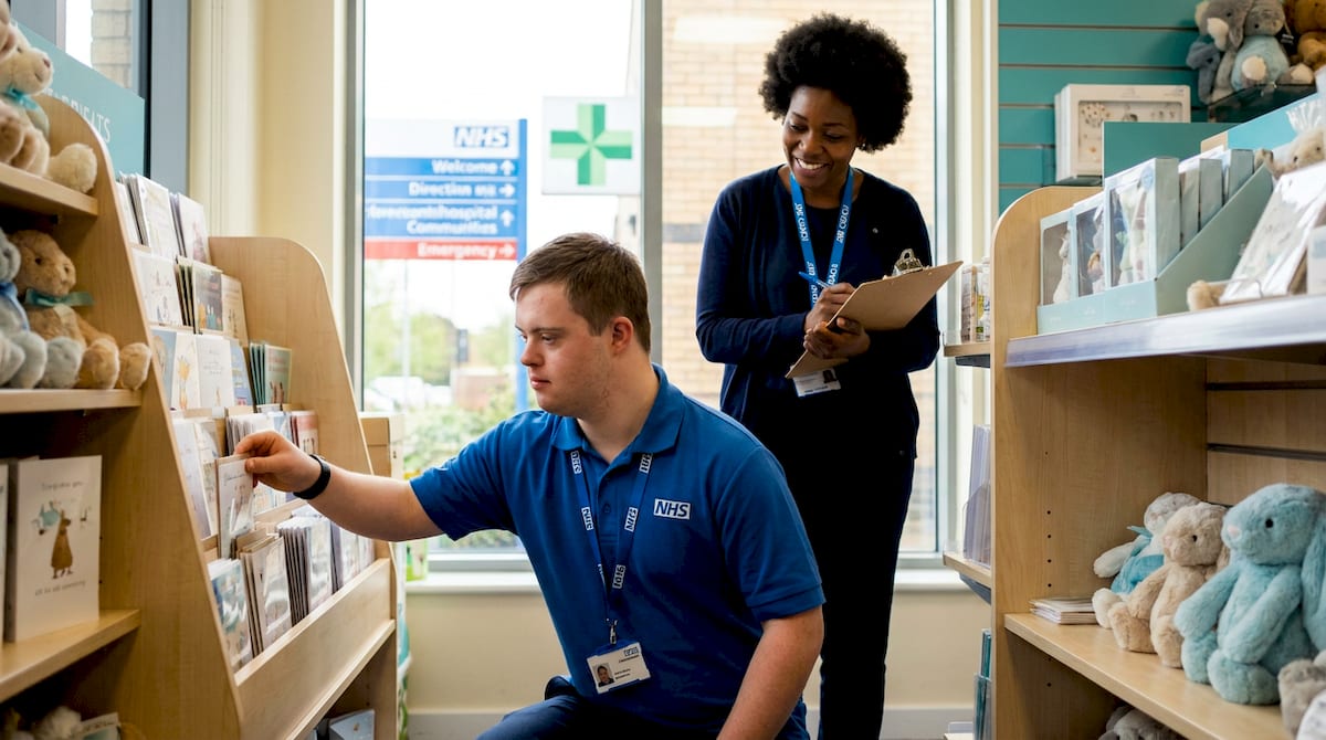 A young man with Down's syndrome in an NHS polo shirt restocks greeting cards in a hospital shop while his job coach watches with a clipboard. AI-generated illustration.